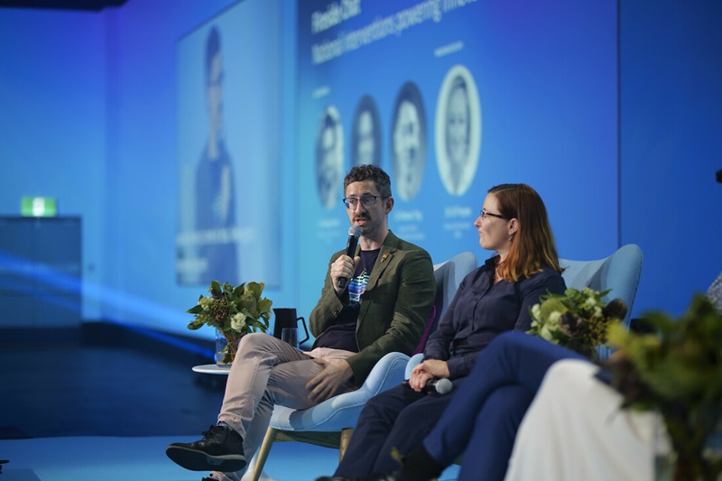 A side angle of two people on a stage at a conference with Dr George Feast seated speaking into a microphone.