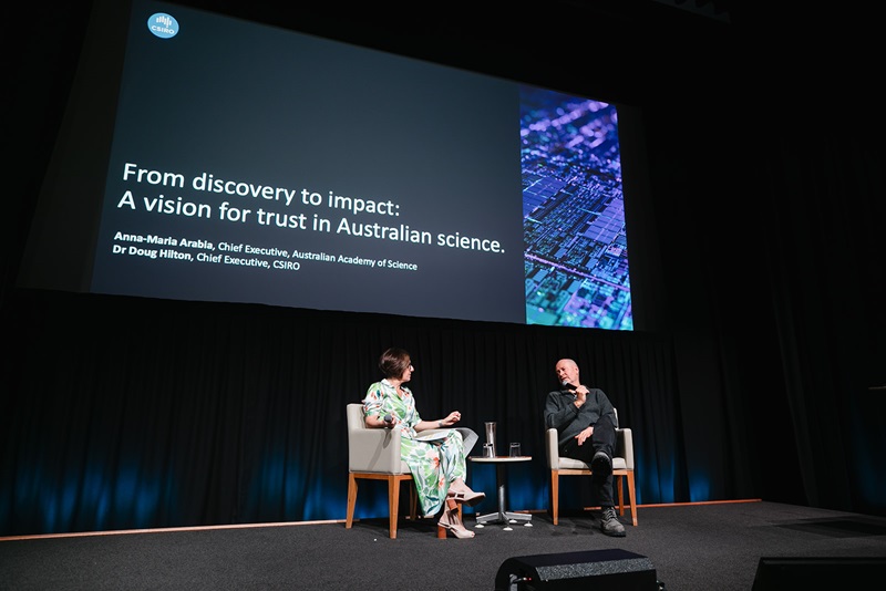 Two people sitting on armchairs with mics talking to one another on a stage. A large backdrop reads 'From discovery to impact: A vision for trust in Australian science'.