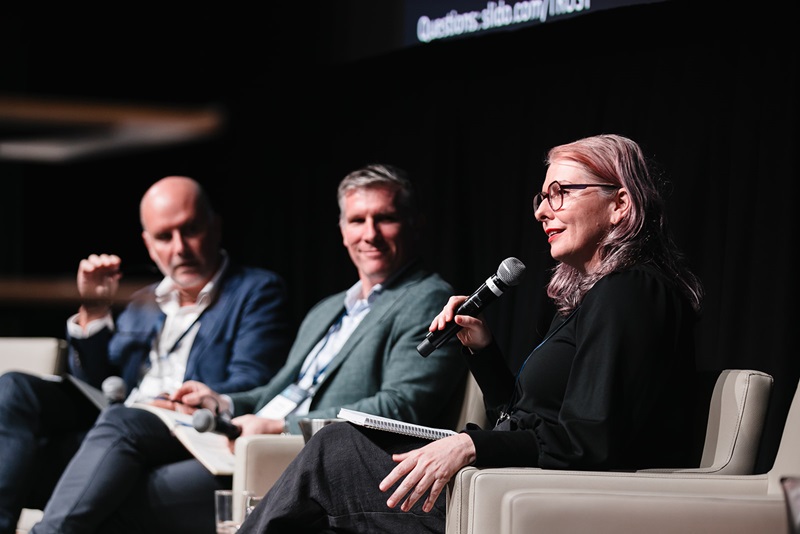 A panel of three seated on a stage with Dr Justine Lacey in the foreground speaking into a microphone.