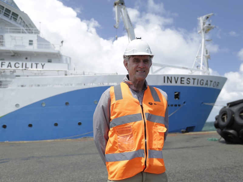 A person in a hard hat and hi vis vest stands on a wharf in front of a blue and white ship.