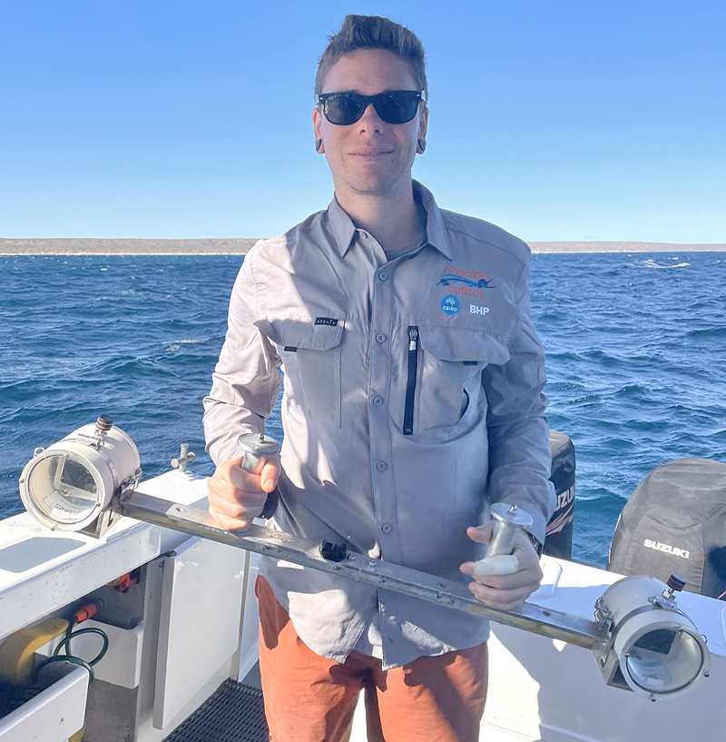Colby standing on the back of a boat holding equipment with sunglasses, sea in the background and land in the distance.