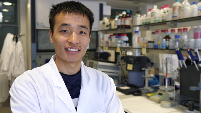 A scientist smiles for the camera whilst wearing a lab coat, in a room full of various scientific instruments and paraphernalia.