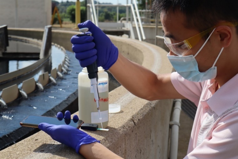A person wearing a face mask and gloves uses scientific equipment and a phone to conduct scientific experiments.