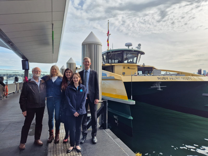 Four people standing in a row on a pier next to a green and yellow Sydney ferry with the name Ruby Payne-Scott painted in white on the ferry.