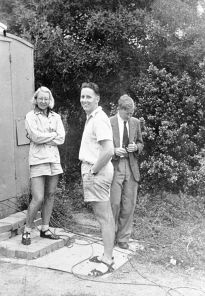 Black and white photo of three people standing outside next to what looks like a garage door, two in shorts facing the camera and one in a suit looking down