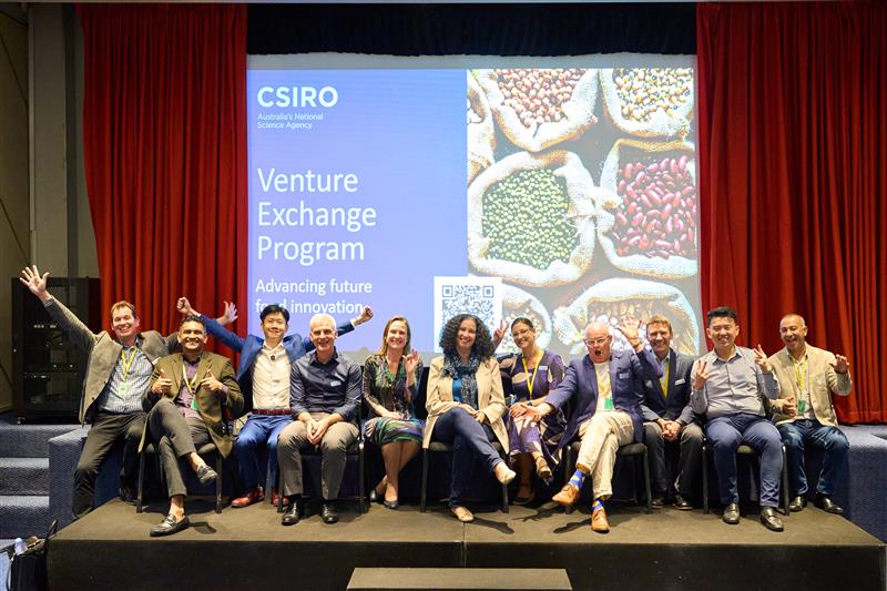 A row of 10-12 people seated on stage facing the camera, a couple to the left with their hands in the air, a large screen behind with images of food and text 'CSIRO Venture Exchange Program'.