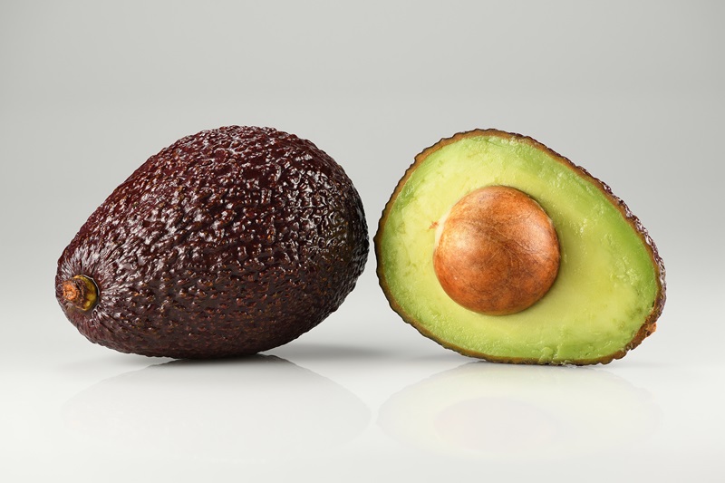 A whole Hass avocado and a halved one showing its seed and creamy green flesh.