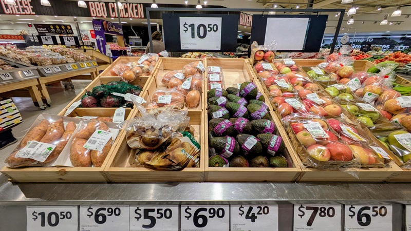 Packaged organic avocados on display in a supermarket alongside apples, onions, and sweet potatoes, with a price tag of $4.20 each.