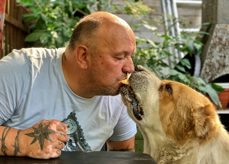 A man and a dog almost touching mouths with two ends of the same bit of food in their mouths. 