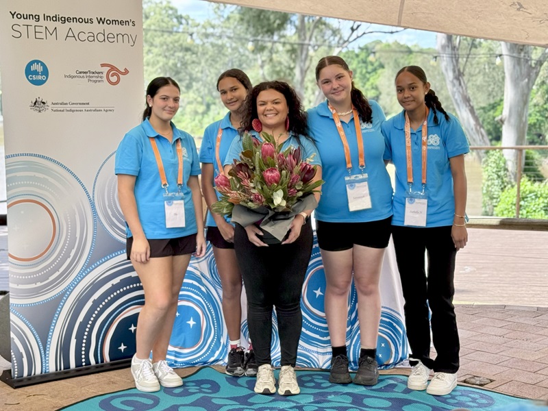Sharnie holding a bunch of flowers with four young students standing behind her, next to a Young Indigenous Womens STEM Academy Program banner.