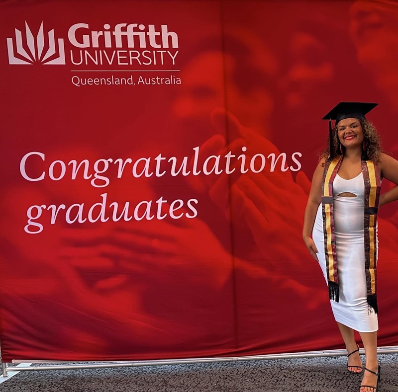 Sharnie in an academic gown and mortar board in front of a Griffith University banner with 'Congratulations graduates'.