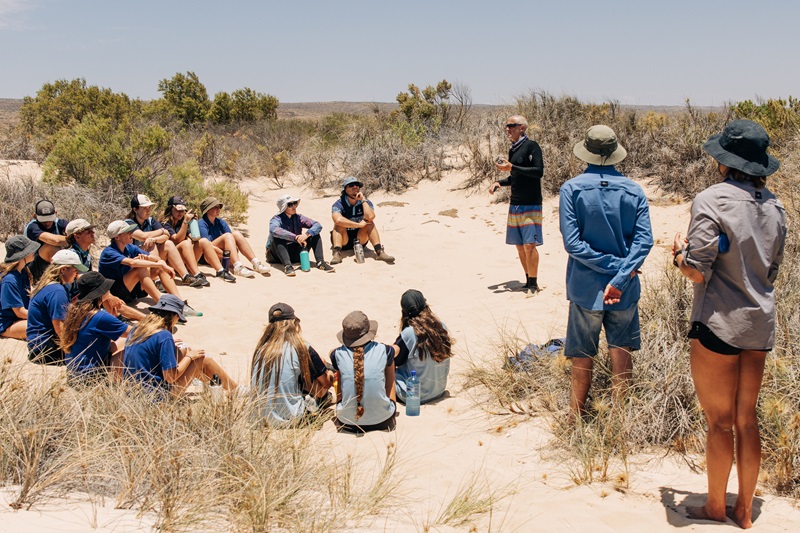 A group of school kids gather on the beach to listen to a scientist. 