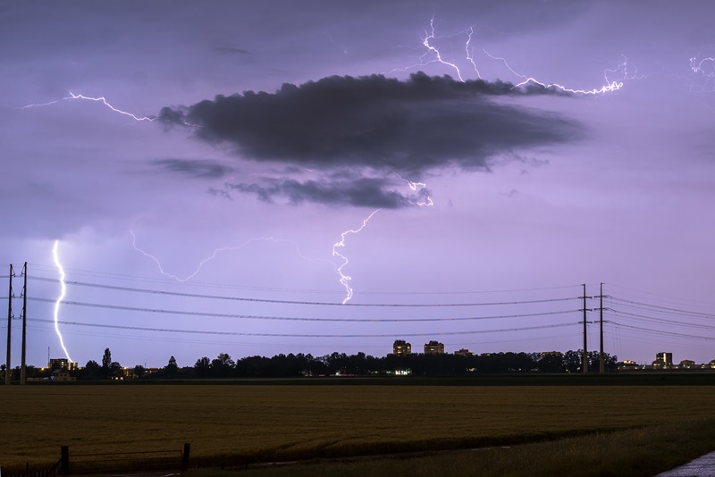 A dark cloud and lightning strikes against a dark puplish sky with transition wires crossing the landscape.