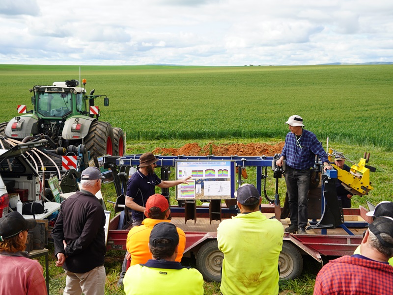 A group of people gathered around a trailer-mounted display with deep ripper components, as presenters from the University of South Australia explain setup options in a green paddock.