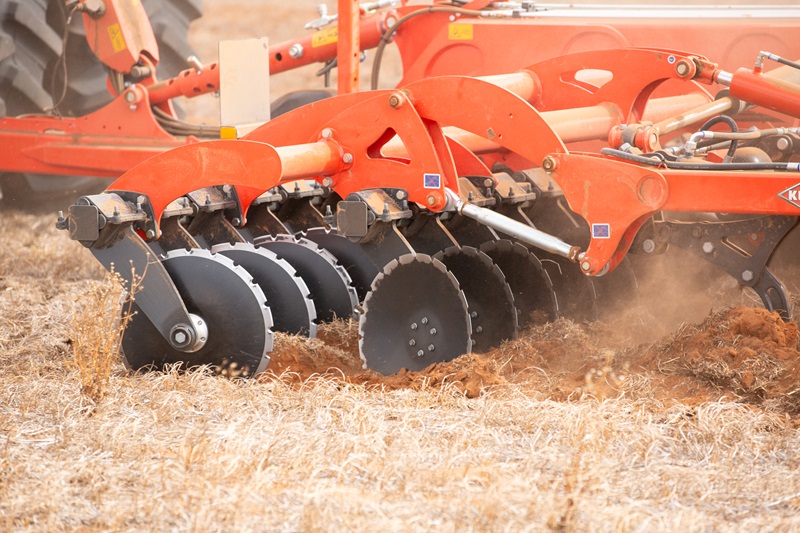 A close-up of a red cultivator with sharp circular blades slicing through dry, sandy soil in a field.