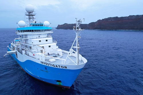 A blue and white ship on the ocean with a volcanic island in the background.