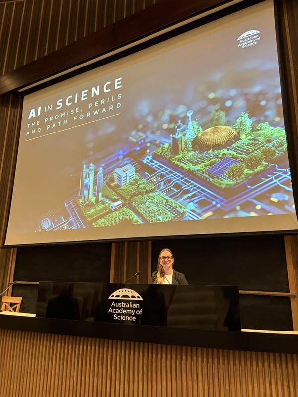 Dr Sarah Hartman standing at a podium beneath a presentation slide reading “AI in Science: The Promise, Perils and Path Forward.”