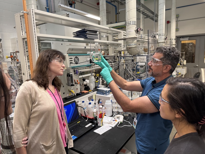 Dia in a lab holding up a glass container with two colleagues looking on