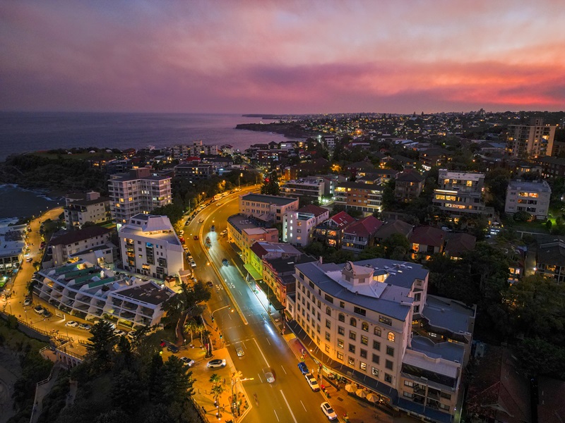 Drone footage of an urban nightscape with orange lights highlighting the road and buildings, and a pink sunset