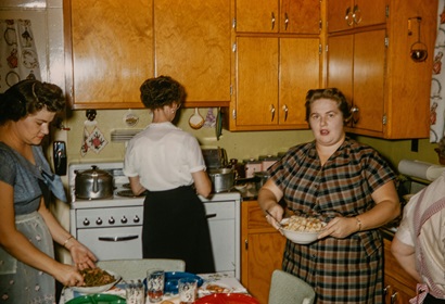 A flashlit photo of three women in a 1940s kitchen preparing a meal, one facing the stove, another looking down at the kitchen table and the third facing the camera.