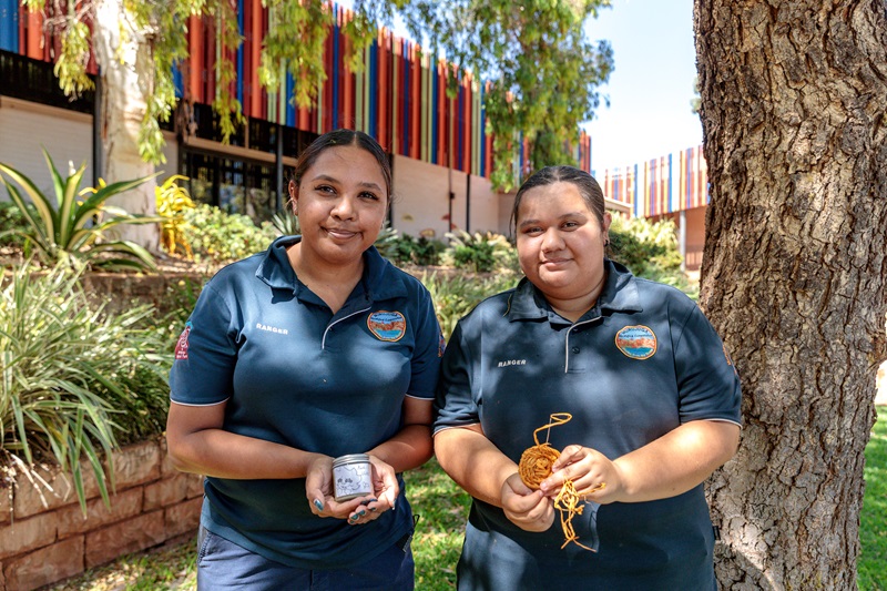 Two women standing in front of a tree holding and jar and weaving example 