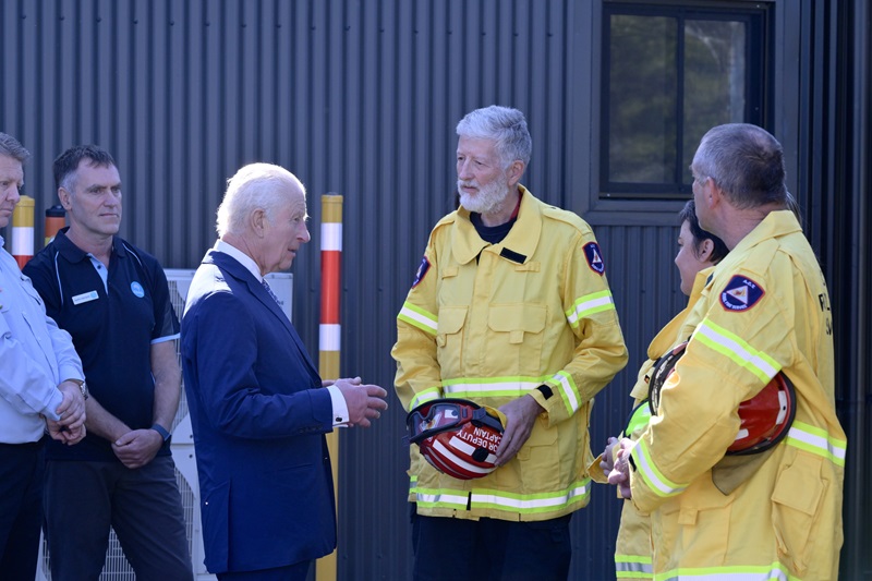 Justin Leonard watches on as firefighters in yellow high viz coats holding their red protective helmets discuss their work with King Charles.