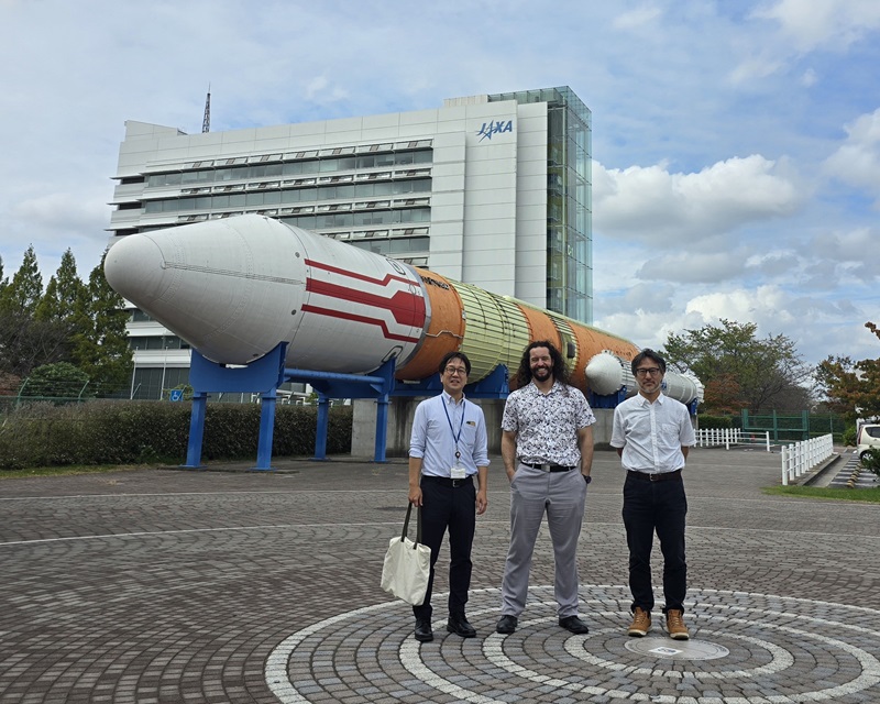 Three researchers standing in front of a large rocket display outside a JAXA building in Japan.