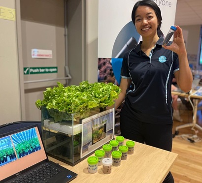 A CSIRO researcher stands beside a small aquaponics display with lush green lettuce growing in a clear tank. She smiles and holds up a vial of material used in the experiment. A laptop and labelled sample jars sit on the table beside her.