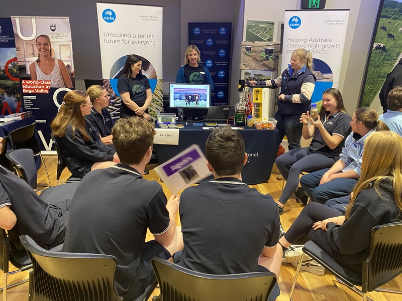 A group of school students sit in front of a CSIRO booth while researchers demonstrate equipment and show examples of algae-based food and protein technologies.