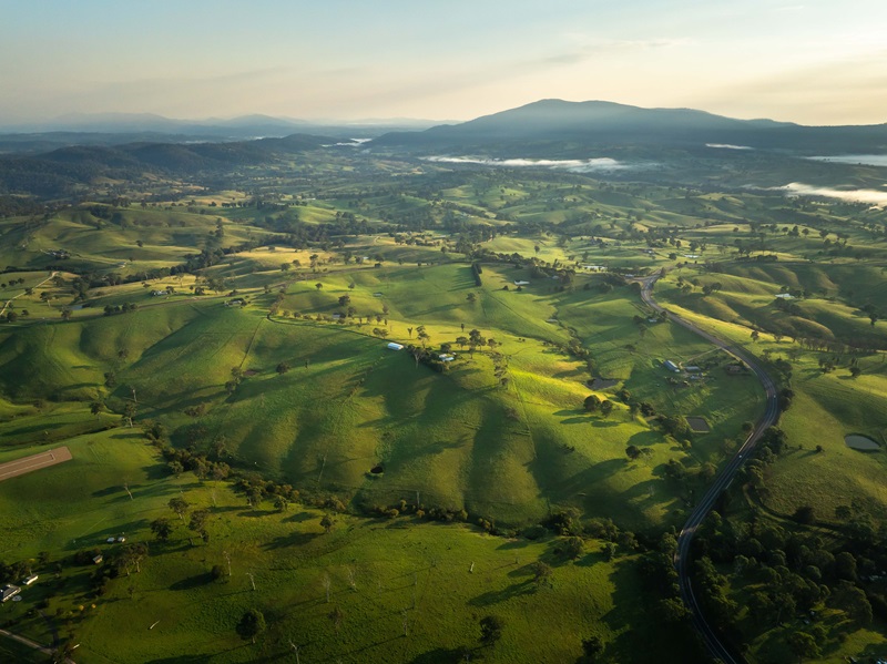 Aerial view of the Bega Valley's rolling green hills at sunrise, with long shadows stretching across the farmland and distant mountains under a light mist.