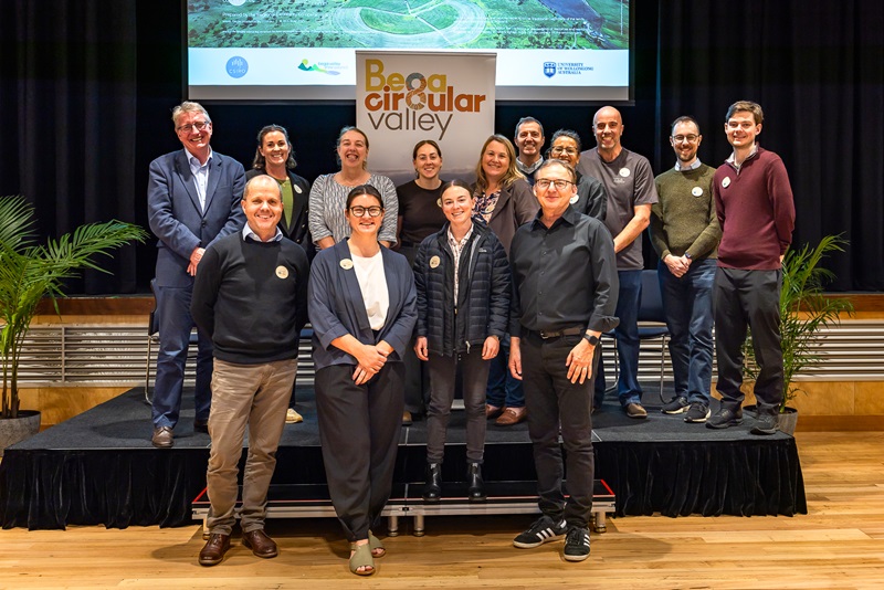 A group of workshop speakers and organisers stand together on stage in front of a Bega Circular Valley banner, smiling for the camera.