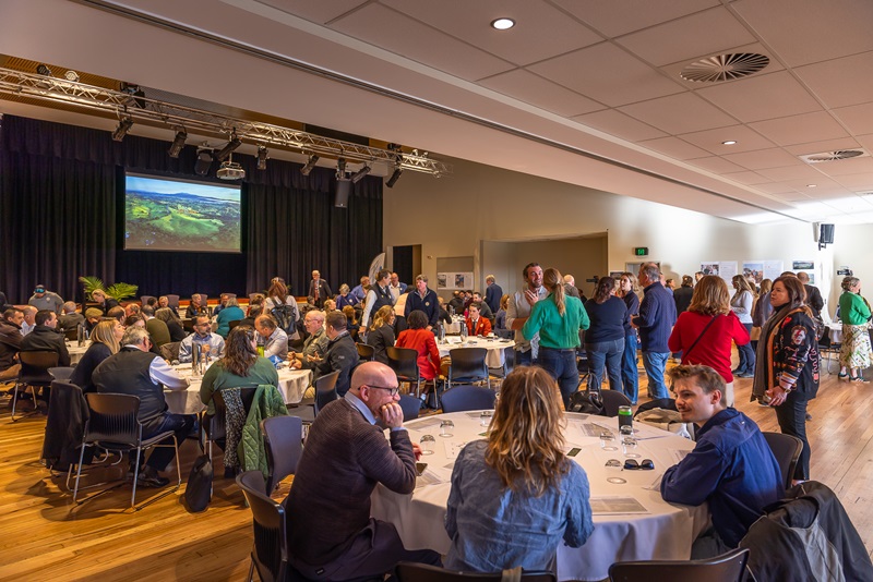 A large community workshop with dozens of participants seated around round tables in a hall, listening, taking notes and discussing circular-economy ideas.