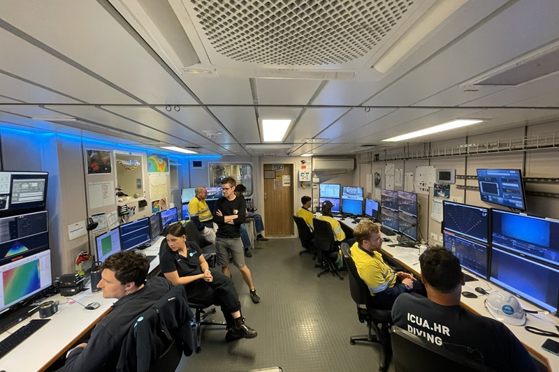 A group of people sit in chairs in front of banks of computer screens in an office.