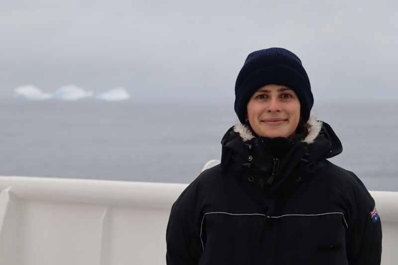 A person in cold weather clothing standing on the deck of a ship with an iceberg on the ocean in the background.