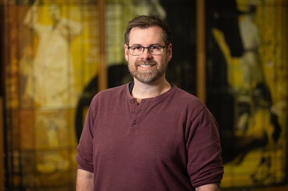 A person wearing a maroon long‑sleeve shirt stands indoors in front of a softly lit background featuring abstract yellow and dark-toned artwork.