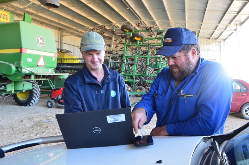 Two men stand beside a vehicle inside a machinery shed, looking at a laptop, with farm equipment visible in the background.