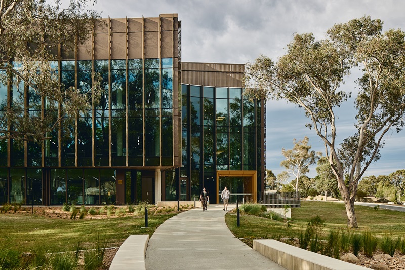A footpath leads to a beautiful building with a glassy exterior. Two people are exiting the yellow doorway of the building, making their way down the path with lawn and Australian native plants on either side. 