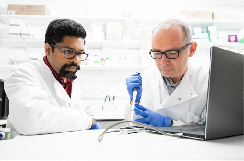 Two scientists in lab coats and glasses handle a pipette, focussed intensely on their work. They are in a clean, white lab with a laptop to the side. 