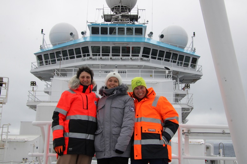 Three people dressed in cold weather gear stand on the bow of a ship with the bridge of the ship above them in the background.