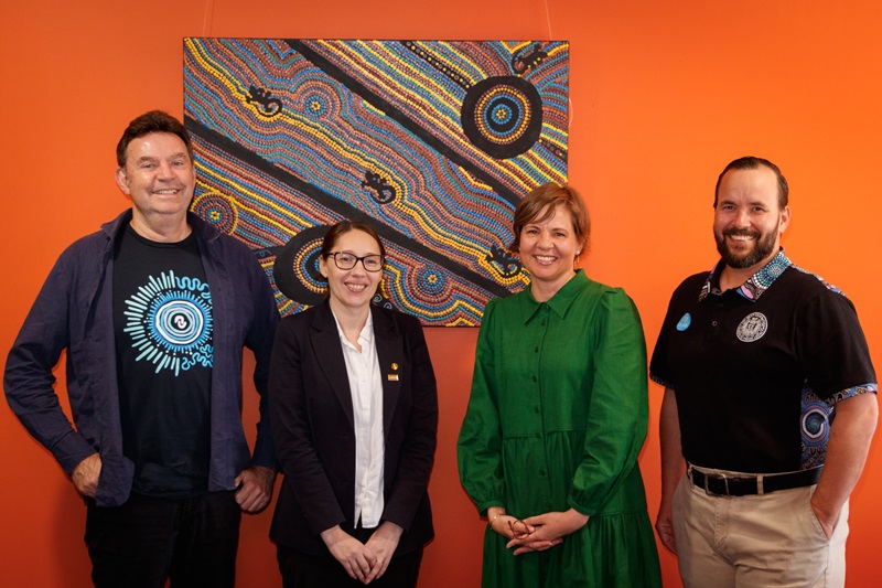 Four people standing in front of an Indigenous painting.