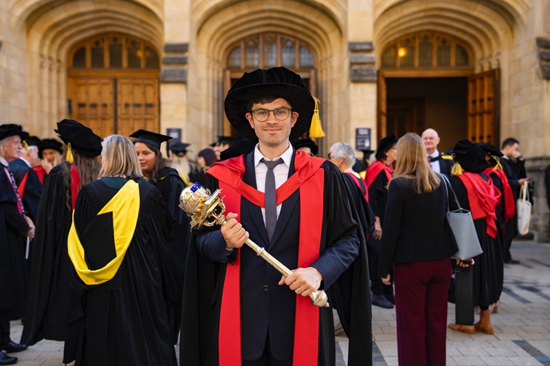 Dr James Manson wearing academic regalia and holding a ceremonial mace at his PhD graduation ceremony.