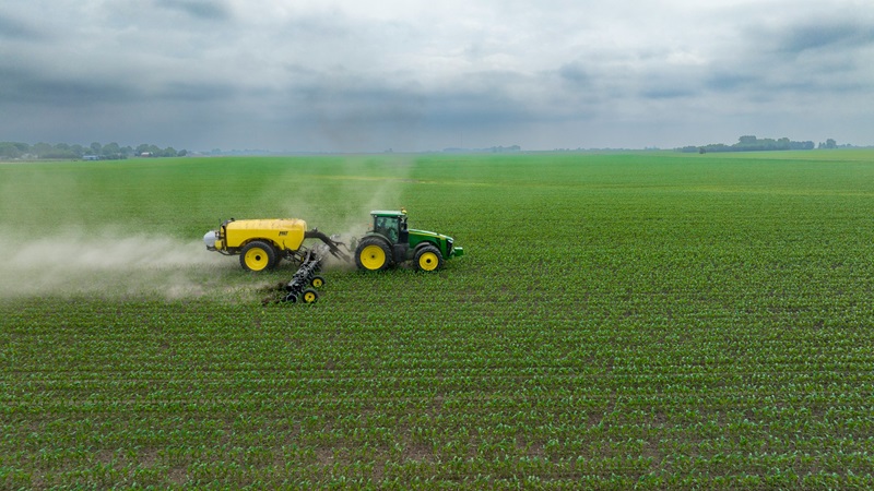 Aerial view of a green and yellow tractor pulling a yellow fertiliser tank across a large green cropping field, with dust rising behind the machinery under an overcast sky.