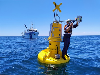 Researcher installs and adjusts a sensor on a yellow ocean monitoring buoy, with a research vessel nearby at sea.
