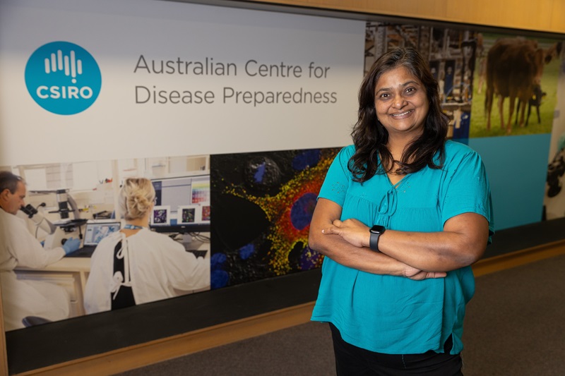 Dr Vidya Bhardwaj stands with arms crossed in front of a wall display featuring the CSIRO logo and the words “Australian Centre for Disease Preparedness”. The display includes images of laboratory research, microscopy and livestock, highlighting the centre’s work in disease preparedness and biosecurity.