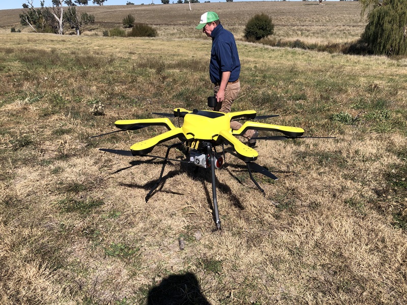 A senior research technician in a green and white cap stands behind a yellow six-rotor drone with a LiDAR sensor slung beneath it, in a dry grass paddock.
