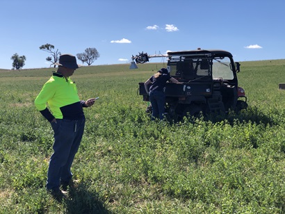 Two researchers, one in a high-visibility shirt, work beside a sensor-equipped utility vehicle in a lush green paddock under a clear sky.