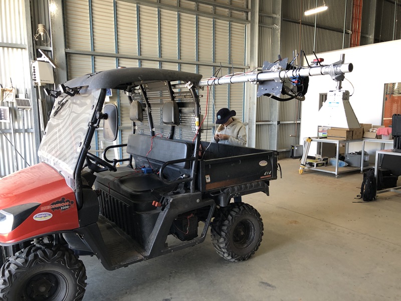 A red and black utility vehicle fitted with a long metal boom carrying sensor equipment, parked in a shed, with a researcher working in its tray.