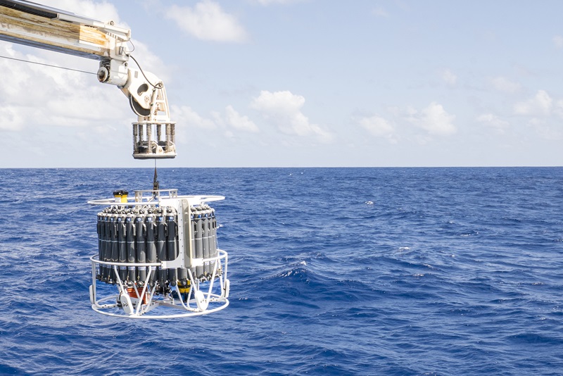 A circular piece of scientific equipment covered in water bottles being lowered into the ocean from a boom.