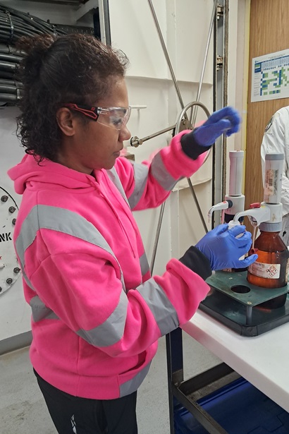 Researcher Sulueti wearing safety glasses, blue protective gloves and a high-visibility pink jacket operates a laboratory dispensing system attached to reagent bottles. She stands beside a laboratory bench in an industrial or research facility, carefully handling equipment used for sample preparation.