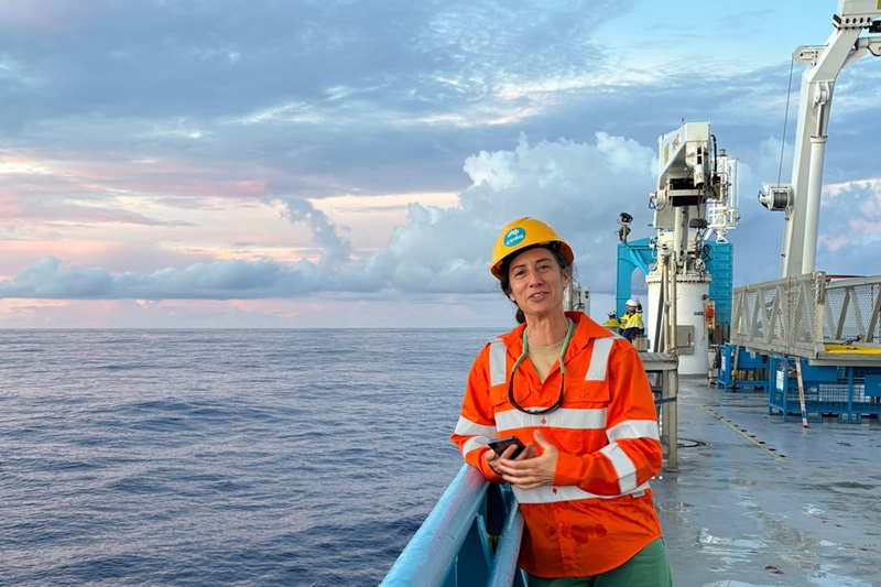 Voyage Chief Scientist Dr Bea Pena-Molino wearing a high-visibility orange safety clothing and a hard hat stands on the deck of Research Vessel Investigator. Scientific equipment and winches are visible on deck, with calm ocean waters and a cloud-filled sky at sunset in the background.